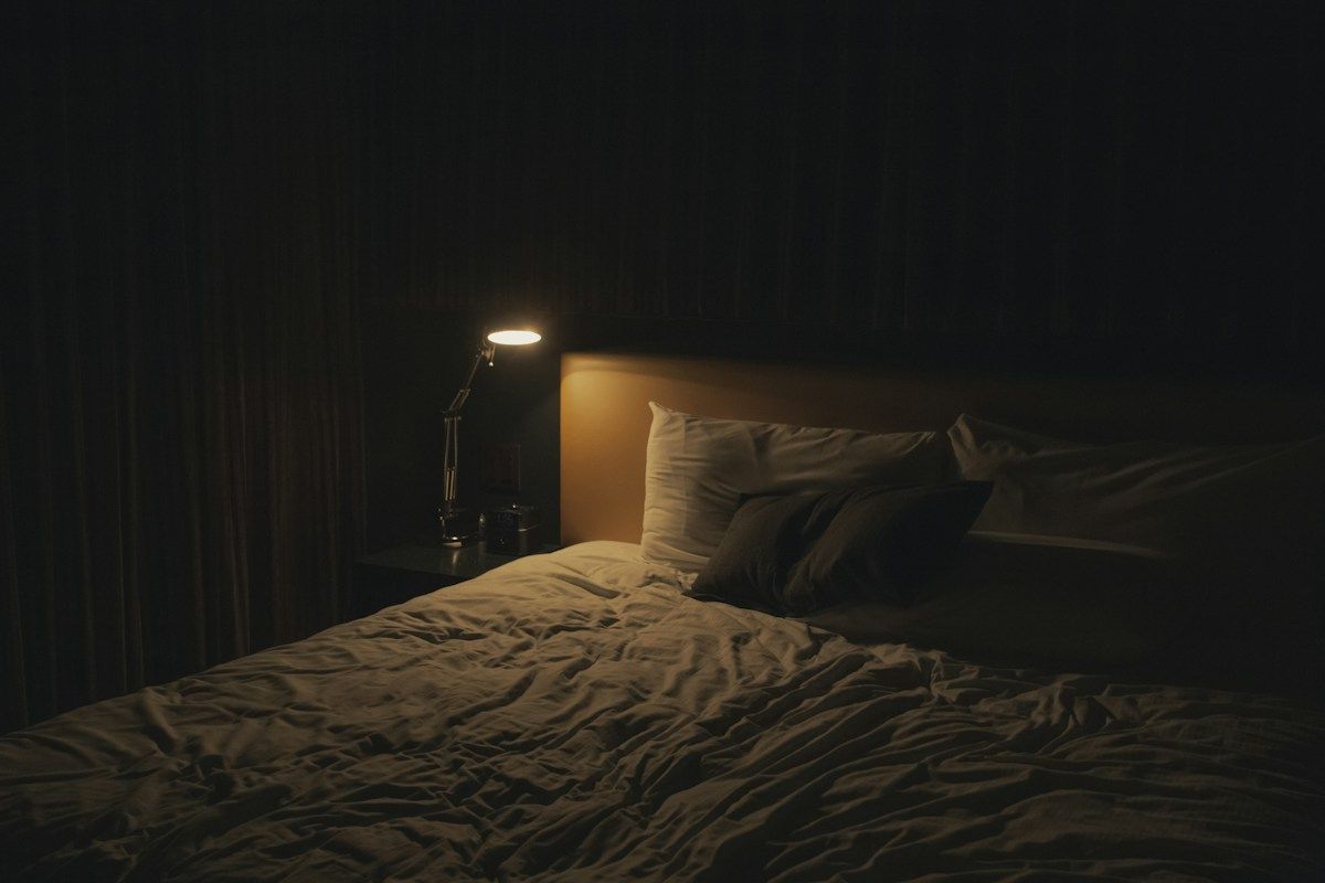 Chambre dans la pénombre, lit défait et petite lampe de lecture